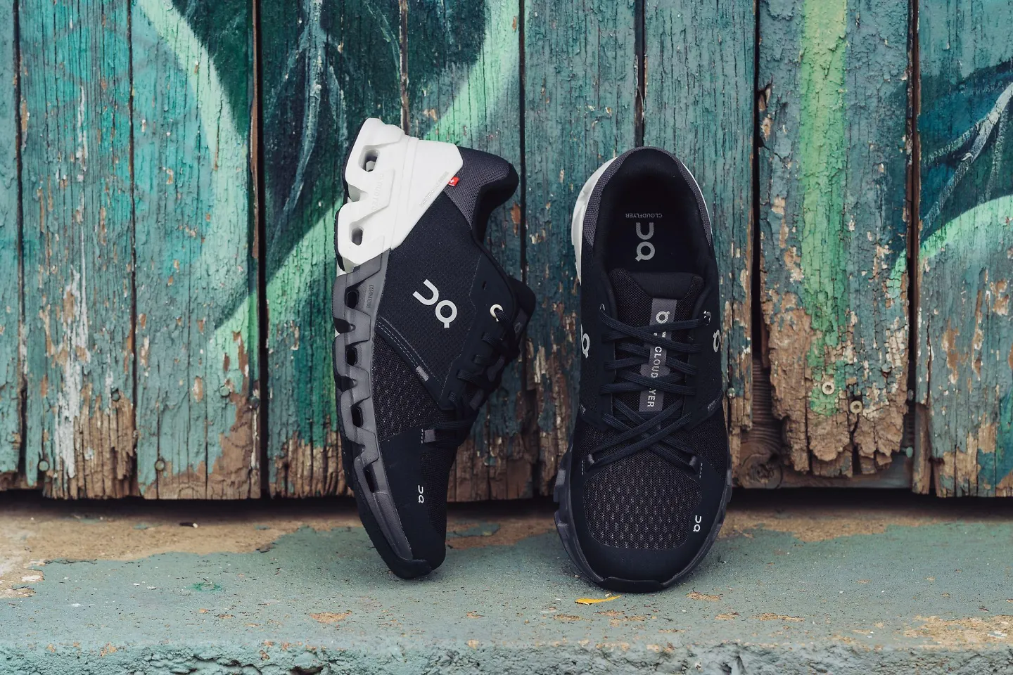 The men's black and white On Cloudflyer 4 sits propped up against a painted blue fence.