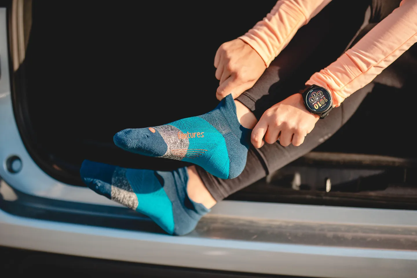 A woman sits on her car, sliding on a pair of Feetures socks.