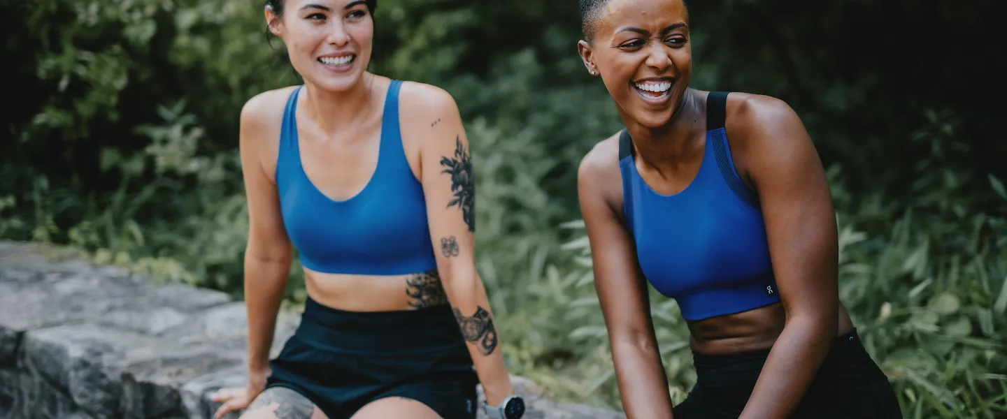 Two women in sports bras sit on a bench laughing.