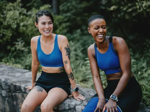 Two women in sports bras sit on a bench laughing.