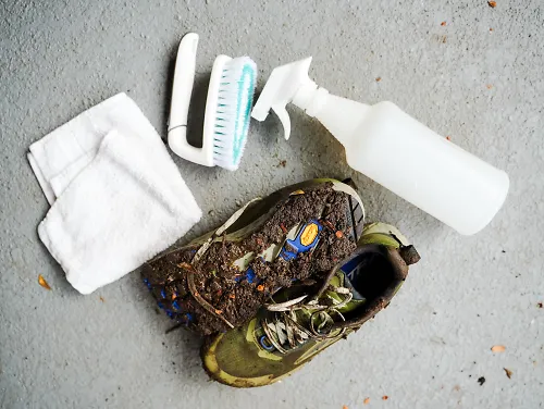 A dirty pair of running shoes lays on the pavement with a cleaning rag, a brush and a spray bottle.