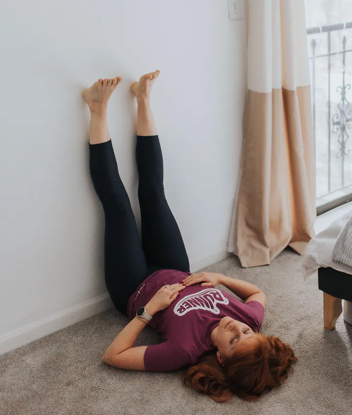 A woman lays on the ground with her feet propped up on the wall.