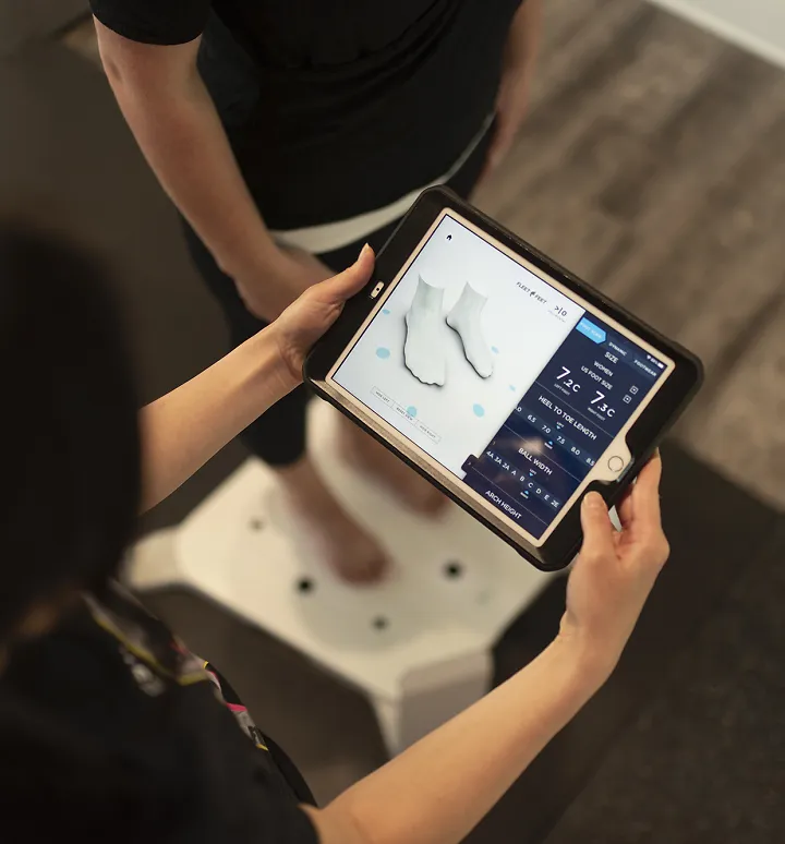 A Fleet Feet outfitter holds an ipad showing a customer's foot scan while the customer stands on the scanner