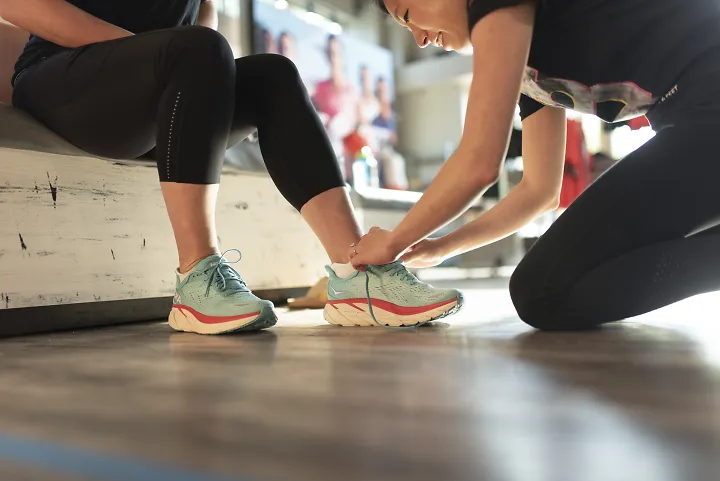 An outfitter crouches on the floor as she laces up a pair of HOKA clifton shoes on her customer's feet. 