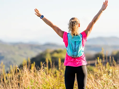 A woman wearing a hydration pack holds up her arms in celebration during a trail run