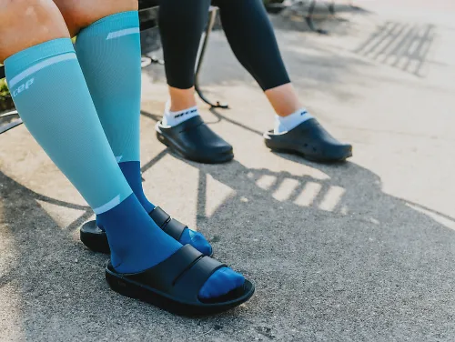 A pictre of two sets of legs, sitting on a bench one wearing OOFOS sandals the other OOCloog. Both have CEP compression socks on with them.