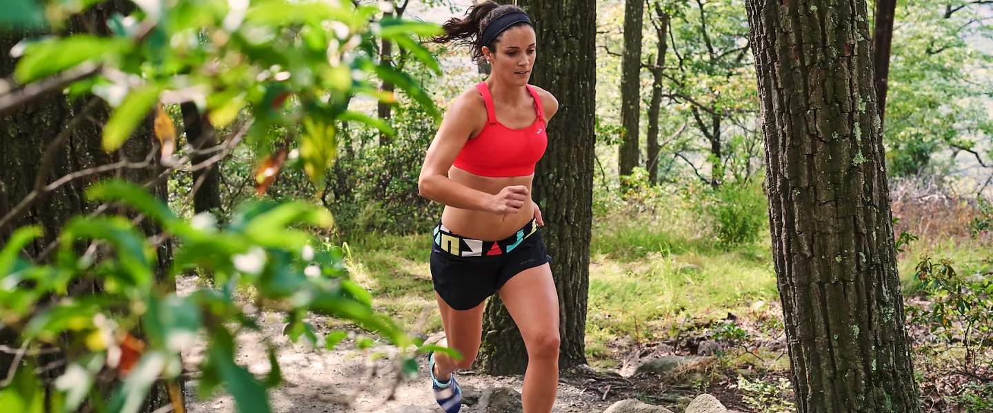 A woman wearing a Brooks Fiona sports bra runs on a trail through the woods