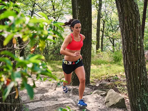 A woman wearing a Brooks Fiona sports bra runs on a trail through the woods