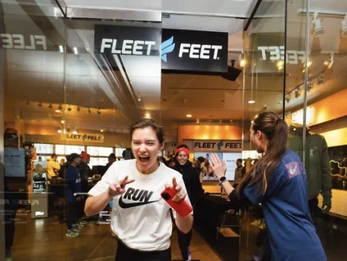A woman in a Nike shirt runs out of a Fleet Feet store looking excited