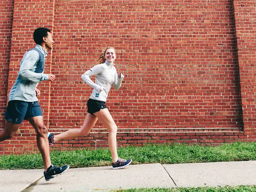 Two people wearing On apparel run past a brick wall